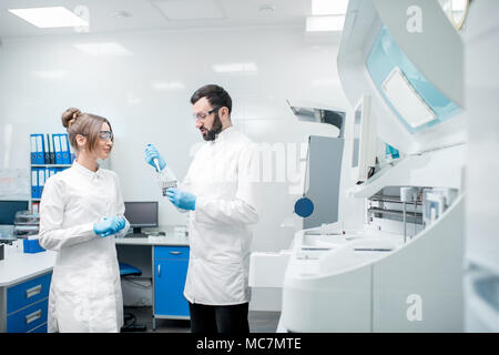 Paar Laboranten in Uniform arbeiten mit Reagenzgläsern stehen in der Nähe der Analyzer Maschine im Labor Stockfoto