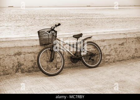 Lonely Fahrrad mit Metall Korb stehend auf betonpfeiler. Seascape Hintergrund mit Fischerbooten auf der Horizontlinie. Getönten Bild. Stockfoto