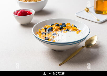 Gesundes Frühstück. Corn Flakes mit Joghurt, Blaubeeren und Chia Samen. Mit Ahornsirup und Himbeeren auf beiger Stoff Hintergrund serviert. Stockfoto