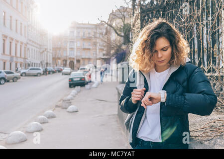 Junge Frau wartet und überprüft, Zeit für Armbanduhren. Redhead curly weibliche steht in der Altstadt in der Nähe der Straße mit Autos schaut auf die Uhr, morni Stockfoto