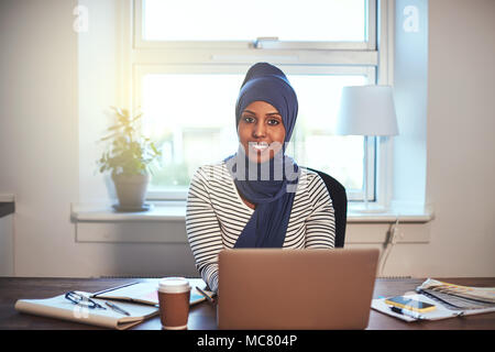 Junge arabische weibliche Unternehmer Tragen eines hijab selbstbewusst lächelnd während der Arbeit auf einem Laptop in Ihrem home office Stockfoto