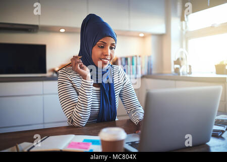 Junge arabische weibliche Unternehmer Tragen eines hijab lächelnd, während an ihrem Küchentisch sitzen, die an einem Notebook arbeitet Stockfoto