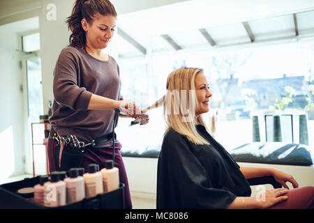 Lächelnde blonde junge Frau sitzt in einem Salon Stuhl in ihr Haar gerichtet, indem ihr Hairstylist Stockfoto