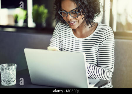 Lächelnden jungen afrikanischen Frau Brille sitzen an einem Tisch das Senden einer SMS-Nachricht auf Ihr Handy und online arbeiten mit einem Laptop Stockfoto