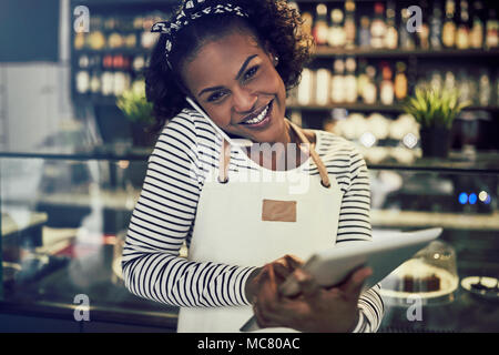Lächelnden jungen afrikanischen Unternehmer Tragen einer Schürze steht in Ihrem Cafe sprechen über ein Handy und das Arbeiten auf einem Tablet Stockfoto
