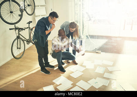 Gruppe von verschiedenen jungen Kollegen stehen im stilvollen modernen Büro lesen gemeinsam ein Dokument von Schreibarbeit verteilen sich auf dem Boden vor o Stockfoto