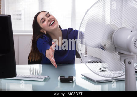 Glückliche junge geschäftsfrau Sitzen auf Stuhl kühlen sich mit elektrischen Ventilator im Büro Stockfoto