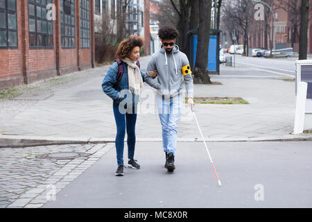 Junge Frau hilft blinden Mann mit weißem Stick beim Überqueren der Straße Stockfoto