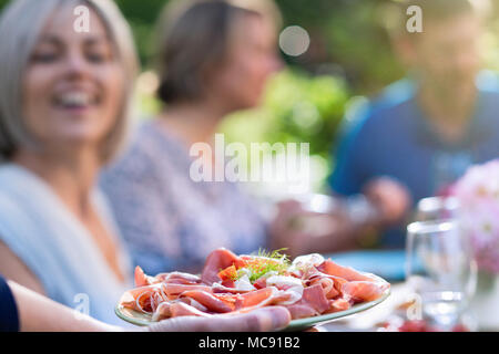 Close-up auf einer Hand eine Schale mit trockenen Schinken und Käse zu Freunden um einen Tisch im Garten versammelt, zusammen Spaß zu haben. Stockfoto
