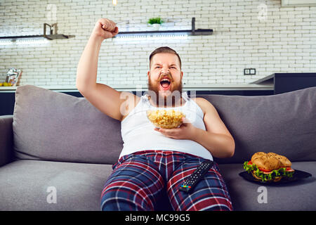 Dick lustig Mann im Schlafanzug einen Burger essen, sitzen auf der Couch, vor dem Fernseher zu Hause. Stockfoto