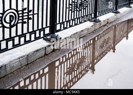 Schwarz cast Gitter von Zaun am Wasser wird in eine Pfütze im verregneten Sommertag wider. Stockfoto