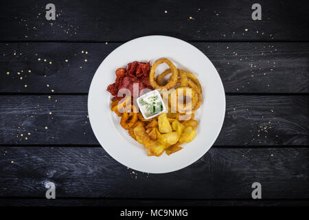 Ringe von Squid, Zwiebelringe, Kartoffelchips, Salami Chips mit Käse Soße auf die weiße Platte mit Holz- Hintergrund. Ansicht von oben. Stockfoto