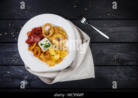 Ringe von Squid, Zwiebelringe, Kartoffelchips, Salami Chips mit Käse Soße auf die weiße Platte mit Bettwäsche Serviette und Gabel, auf der hölzernen backgrou Stockfoto