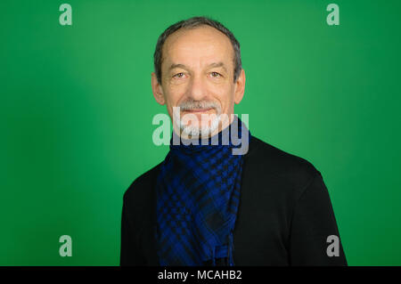 Gut aussehende gut Aussehender älterer grauhaariger Mann mit Gut getrimmten Bart in schwarzen Pullover und blau karierten Schal um den Hals in die Kamera schaut auf grünem Hintergrund im Studio Stockfoto