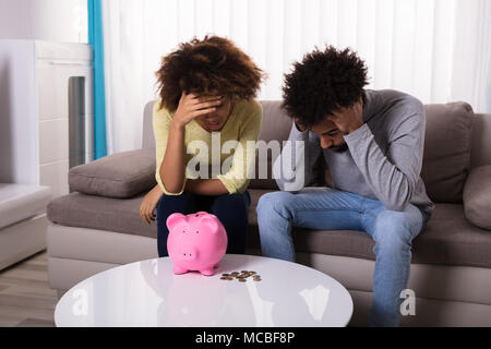 Traurig Paar sitzt auf einem Sofa mit Sparschwein und Münzen auf Tisch Stockfoto