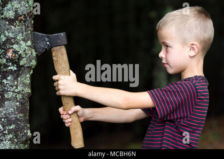 Kleine Jungen mit schweren alten Bügeleisen ax schneiden Baum Trumpf in Wald auf Sommertag. Outdoor Aktivitäten und körperliche Arbeit. Stockfoto