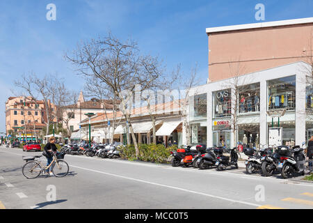 Frau treibt ein Fahrrad in Granviale Santa Maria Elisabetta, Lido, Venedig, Venetien, Italien mit geparkte Motorräder vor den Geschäften und Restaurants. Bik Stockfoto