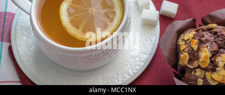Tee mit Zitrone in einem weißen Schale auf eine Untertasse mit Würfeln von raffinated Zucker und Schokolade Muffin mit Pistazien auf einem roten Serviette. Hohe Betrachtungswinkel, panora Stockfoto