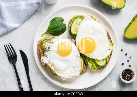 Zwei Toasts mit sunny side up Ei, Avocado und Gurke auf weiße Platte, Ansicht von oben. Gesundes Frühstück Stockfoto