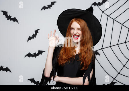 Halloween Hexe Konzept - Happy Halloween Hexe Ingwer Haar halten ok Zeichen mit den Fingern über grauer Hintergrund posiert. Stockfoto