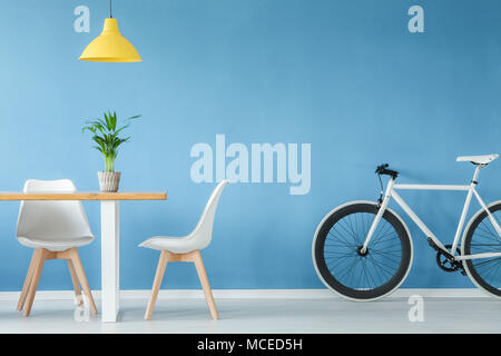 Minimal, modernes Interieur mit zwei Stühlen, ein Fahrrad, eine Tabelle mit einer Anlage und eine gelbe Lampe oben, gegen die blaue Wand Stockfoto