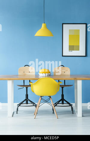 Minimal Style symmetrische Interieur mit einem gelben Lampe hängt über einem Holztisch mit einer Melone in eine Schüssel, Stühle und ein Poster auf einer blauen Wand Stockfoto