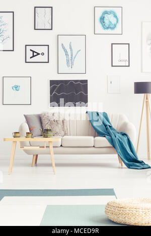 Blaue Decke auf beige Sofa gegen weiße Wand mit Plakaten im Wohnzimmer Interieur mit Pouf Stockfoto