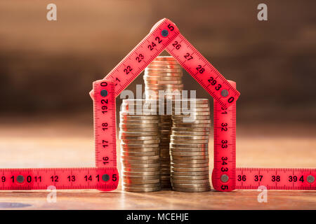 Stapel von Münzen unter dem Haus mit roten Maßband auf dem Schreibtisch aus Holz Stockfoto