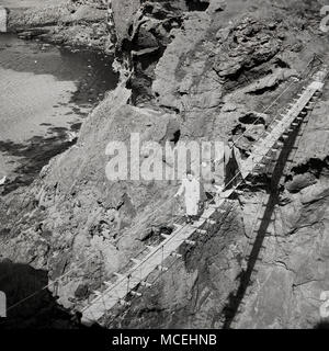 1950, historische, ein Mann und eine Frau, die über die Carrick-a-Rede rope bridge in der Nähe von Ballintoy, Co Antrim, Nordirland. 30 Meter über dem felsigen Klippen, die Brücke verbindet das Festland zu der winzigen Insel Carrickarede und ist in Irland eindeutig. Stockfoto