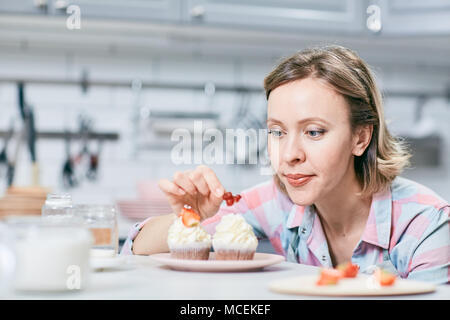 Portrait von Blond kaukasischen Frauen kochen Dekorieren lecker cream Cupcakes mit frischen Beeren in der Küche Stockfoto