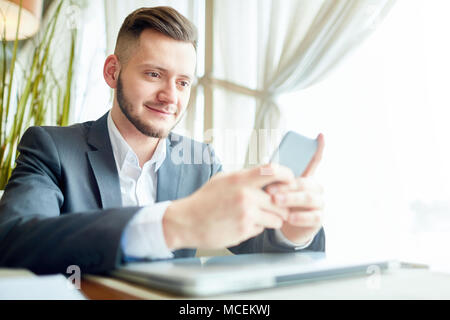 Low Angle View von stattlichen Kaukasischen manager SMS-Nachrichten auf dem Smartphone und lächelnd während der Sitzung in Cafe auf Mittagspause Stockfoto