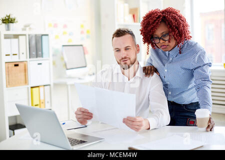 Rothaarige African American woman reading Papier mit ihren männlichen Kaukasischen Kollegen sitzen am Schreibtisch in modernen Büro Stockfoto