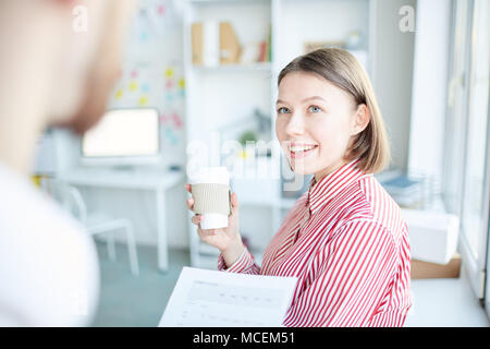 Junge schöne weibliche Büroangestellte Chatten mit Kollegen, Kaffee zum Mitnehmen und freundlich lächelnd Stockfoto