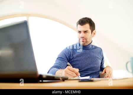 Portrait von Casual jungen Mann mit Rollkragen Pullover und Suchen thoughtul. Arbeiten am Laptop während des Schreibens mit Stift Dokumente. Home Office. Stockfoto