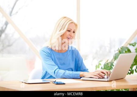 Lächelnd im mittleren Alter Geschäftsfrau Tippen auf Laptop, während am Schreibtisch sitzen und arbeiten von zu Hause aus. Home Office. Stockfoto