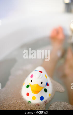Nahaufnahme der bunt getupft Quietscheentchen in der Badewanne mit Blasen und Beine von jemand sichtbar unter Wasser im Hintergrund Stockfoto