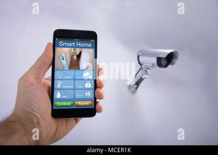 Die menschliche Hand mit Smart Home Anwendungen auf dem Smartphone in der Nähe von CCTV-Kamera an der Wand Stockfoto