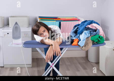 Unglückliche Frau Schiefen auf dem Bügelbrett Neben Stapel von Kleidung Stockfoto