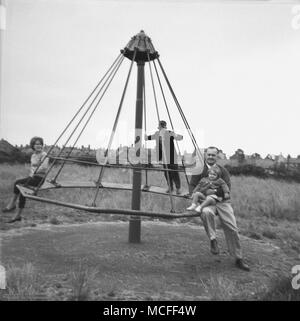 Liverpool, Großbritannien, 1960er Jahre, nach der Schule, Bild zeigt ein Vater mit seinen Kindern im Teenageralter und halten ein Kind sitzt auf einem Metall Merry-go-round an einem alten Spielplatz im Freien. Stockfoto