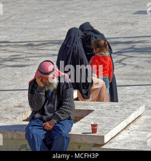 Amman, Jordanien, 11. März 2018: eine traditionell gekleidete Bedouin sitzt auf einer Bank auf Stein und Gespräche am Telefon, während auf der anderen Seite zwei Frauen in b Stockfoto