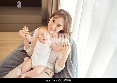 Eine Mutter mit einem Baby auf dem Arm sitzt auf einem Stuhl vor einem Fenster im Zimmer. Stockfoto