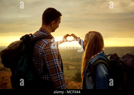 Ein paar Touristen mit Rucksäcken, ein Symbol für die Herzen, mit den Händen auf die Natur am Abend bei Sonnenuntergang. Ansicht von hinten. Stockfoto