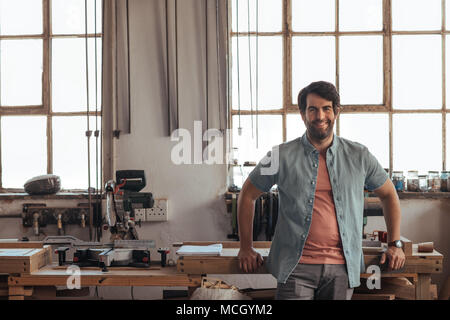 Portrait einer jungen woodworker lächelnd souverän und lehnte sich gegen eine Werkbank voller Werkzeuge in seiner Schreinerei Stockfoto