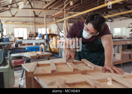 Qualifizierte junge Tischler tragen Schutzbrille und Maske hand Schleifen von Holz, während allein in seinem großen holzwerkstatt arbeiten Stockfoto