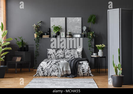 Graue Decke auf gemusterte Bettwäsche in floralen Schlafzimmer Einrichtung mit Pflanzen auf Regal und Gemälde Stockfoto
