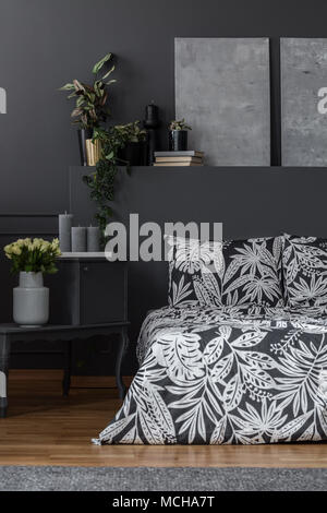 Graue Gemälde auf dunklen Wand über gemusterte Bettwäsche in dunklen Schlafzimmer Innenraum mit Blumen Stockfoto
