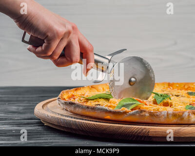 Stahl pazza Cutter. Schneiden leckeres italienisches Gericht auf Holz Stativ isoliert. Woman's Hand mit einem Messer schneiden die Pizza auf weißem Hintergrund close-up Stockfoto