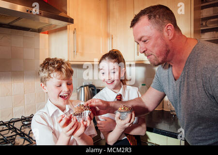 Zwei Kinder sind in der Küche ihres Hauses nach der Schule. Sie essen Cupcakes und der Vater stiehlt einige Tüpfelchen oben von seinen Söhnen cak Stockfoto