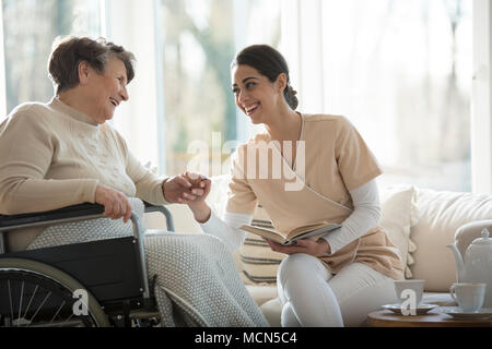 Senior in einem Rollstuhl und schöne Krankenschwester und lächelnd die Hände beim Lesen eines Buches Stockfoto