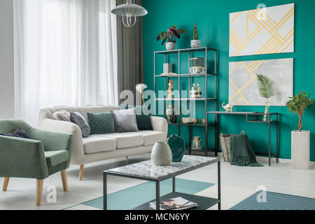 Helles Sofa mit vielen Kissen neben einem grünen Sessel im Wohnzimmer Interieur mit Metall Möbel und geometrischen Gemälde Stockfoto